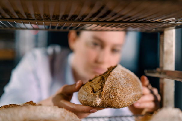 приваблива жінка-пекарня між полицями виглядає і перевіряє свіжоспечений хліб дуже ретельно хлібопекарської промисловості - Фото, зображення