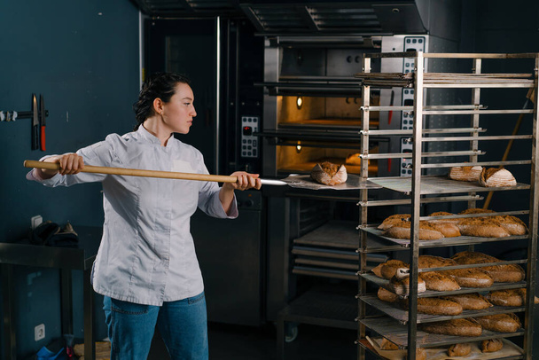 panadero femenino saca pan fresco recién horneado del horno y lo pone en el estante en la cocina de la panadería profesión culinaria - Foto, imagen