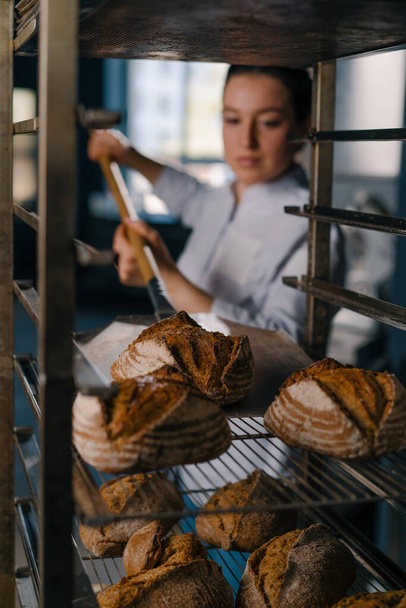 女性のパン屋は,オーブンから新鮮なパンを取り出し,パン屋の料理の専門家のキッチンに棚に置きます - 写真・画像