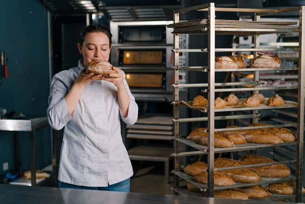 attractive female baker takes freshly baked fragrant bread from the shelf sniffs it checks quality bakery professional kitchen baking - Fotografie, Obrázek