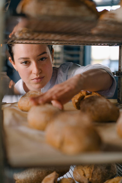 atraktivní pekařka mezi regály hledá a kontroluje čerstvě upečený chléb velmi pečlivě pekařský průmysl - Fotografie, Obrázek