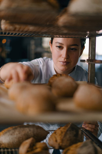 atractiva hembra panadero entre estantes buscando y comprobando pan recién horneado muy cuidadosamente industria panadería - Foto, imagen
