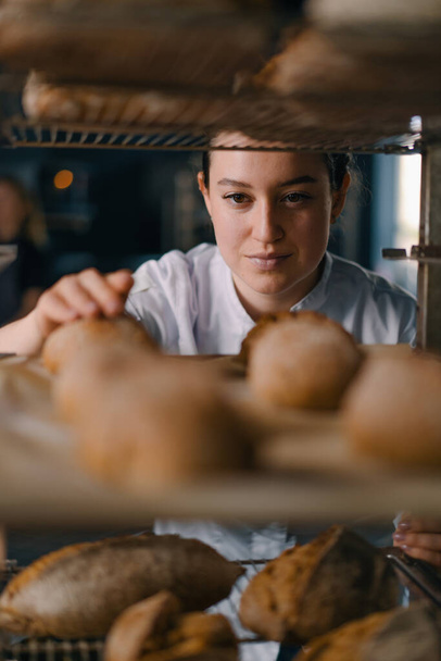 attractive female baker between shelves looking and checking freshly baked bread very carefully bakery industry - Photo, Image