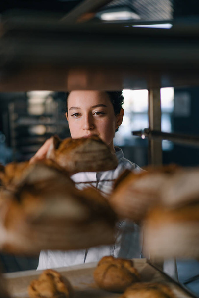 приваблива жінка-пекарня між полицями виглядає і перевіряє свіжоспечений хліб дуже ретельно хлібопекарської промисловості - Фото, зображення
