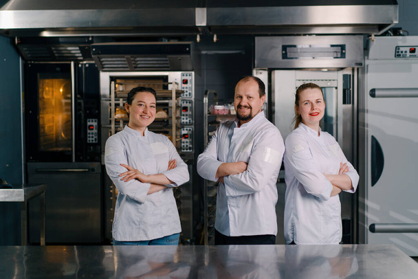 zespół uśmiechniętych profesjonalnych piekarzy w profesjonalnej kuchni w piekarni przed rozpoczęciem produkcji piekarskiej - Zdjęcie, obraz