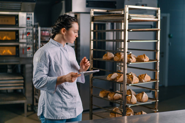 kobieta piekarz w mundurze w pobliżu półek ze świeżo upieczonym chlebem trzyma tabliczkę w rękach sprawdza piekarnię chleba - Zdjęcie, obraz