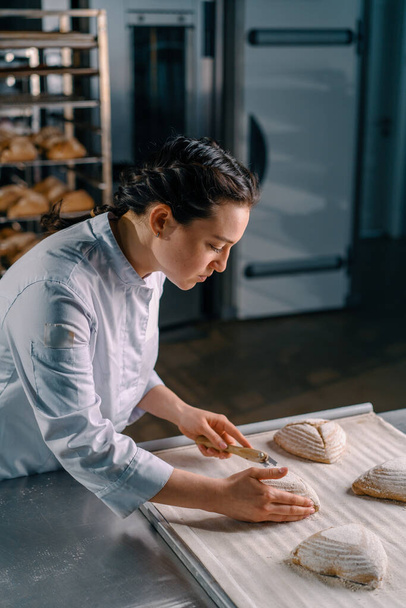 belle femme boulangerie coupes modèles avec un couteau de boulangerie professionnelle sur pain cru petits pains avant la cuisson pâtisseries de production de boulangerie - Photo, image