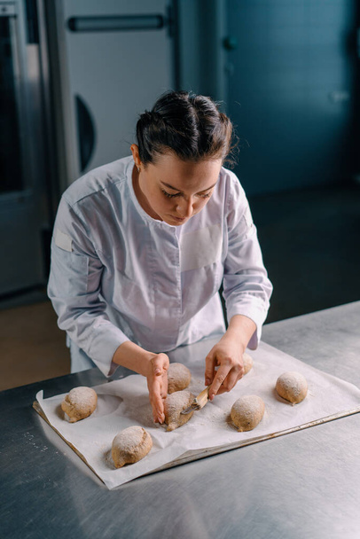 piękna kobieta piekarz tnie wzory z profesjonalnym nożem piekarskim na bułki surowego chleba przed pieczeniem ciasta piekarniczego produkcji - Zdjęcie, obraz