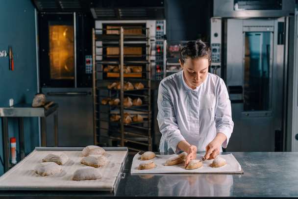 krásná žena pekař řeže vzory s profesionálním pekařským nožem na syrové chlebové buchty před pečením pekařské výroby pečiva - Fotografie, Obrázek