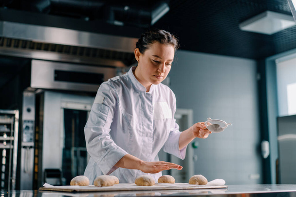 συμπυκνωμένη όμορφη γυναίκα αρτοποιός πασπαλίζει αλεύρι μέσω κόσκινο ακατέργαστο ψωμί φρέσκια ζύμη πριν από το ψήσιμο αρτοποιείο παραγωγής - Φωτογραφία, εικόνα