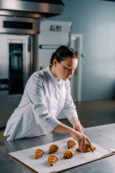beautiful woman baker cuts ready freshly baked hot crispy aromatic croissants with a knife and checks dough bakery production - Foto, Imagem