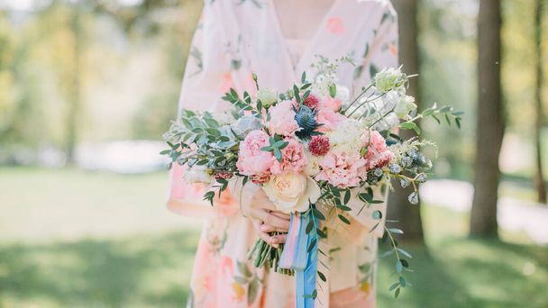 Una novia con una bata rosa en el jardín tiene un ramo de bodas con equeveria, peonías y rosas rosadas y blancas. Cintas azules y rosas cuelgan del ramo. - Foto, imagen