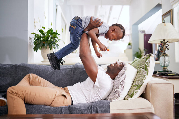 Familia negra, avión y padre con hijo en un sofá con amor, levantando y jugando en su casa juntos. Volar, niño y diversión con los padres en el sofá, reír y jugar, vinculación y juegos en la sala de estar. - Foto, imagen