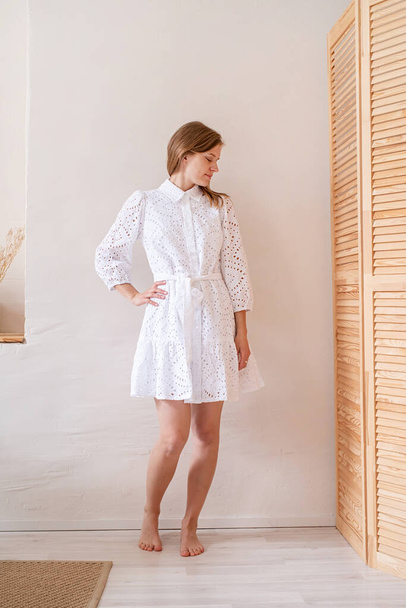 Beautiful caucasian woman in white summer fashion dress standing barefoot on white wall background, looking away. Boho or scandi interior - Foto, Imagen