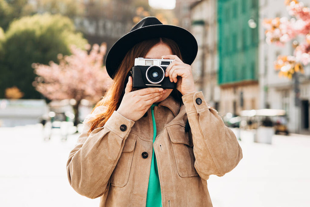 カメラで彼女の顔を覆う写真家の肖像。通りの背景にポーズバックパック付きのケアフリー女性。積極的な観光客屋外。陽気な30代の女の子で帽子あります探検都市 - 写真・画像
