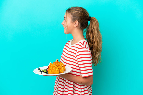 Маленька біла дівчинка тримає вафлі ізольовані на синьому фоні, сміючись у бічному положенні
 - Фото, зображення