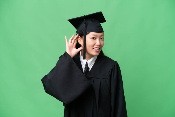 Young university graduate Asian woman over isolated background listening to something by putting hand on the ear - Foto, Imagem