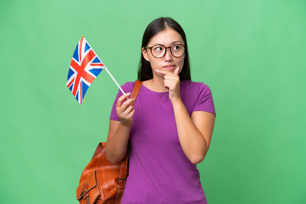 Young Asian woman holding an United Kingdom flag over isolated background having doubts and thinking - Photo, Image