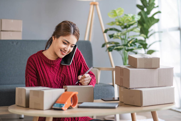 A portrait of a young Asian woman, e-commerce employee sitting in the office full of packages in the background write note of orders use a calculator and smartphone for SME business ecommerce and - Фото, зображення