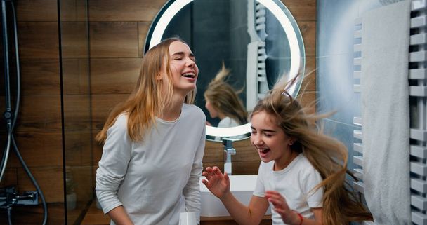Two funny Caucasian girls with beautiful long hair dancing and singing with hair dryer in modern bathroom at home. - Φωτογραφία, εικόνα