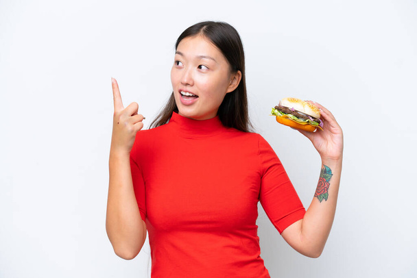 Junge Asiatin hält einen Burger isoliert auf weißem Hintergrund in der Absicht, die Lösung zu erkennen, während sie einen Finger hebt - Foto, Bild
