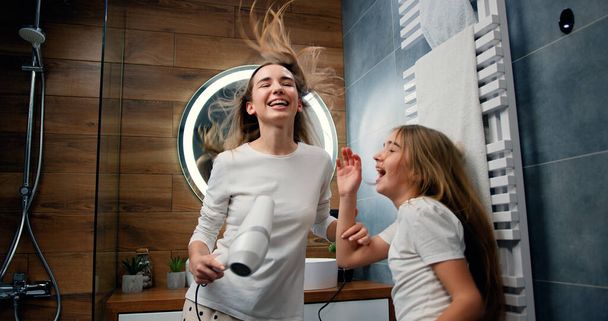 Funny young woman and 10 years old girl with long hair in pajamas drying her voluminous hair by hair dryer while dancing laughing and looking at each other in modern bathroom in home - Photo, image