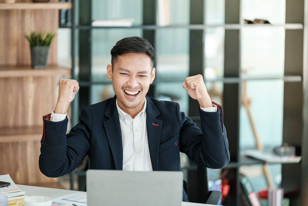 Successful and happy young Asian man, businessman, happy, shows with hand yes. Made a contract. Won money. Sitting with a smart phone or notebook at the table in a modern office. High quality photo - Fotografie, Obrázek