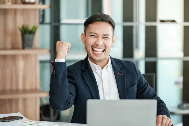 Successful and happy young Asian man, businessman, happy, shows with hand yes. Made a contract. Won money. Sitting with a smart phone or notebook at the table in a modern office. High quality photo - 写真・画像