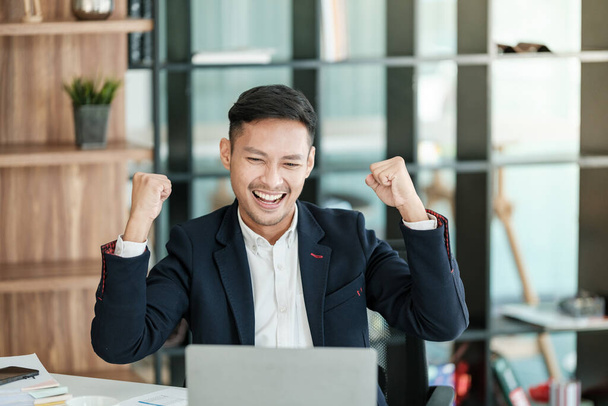Successful and happy young Asian man, businessman, happy, shows with hand yes. Made a contract. Won money. Sitting with a smart phone or notebook at the table in a modern office. High quality photo - Fotografie, Obrázek