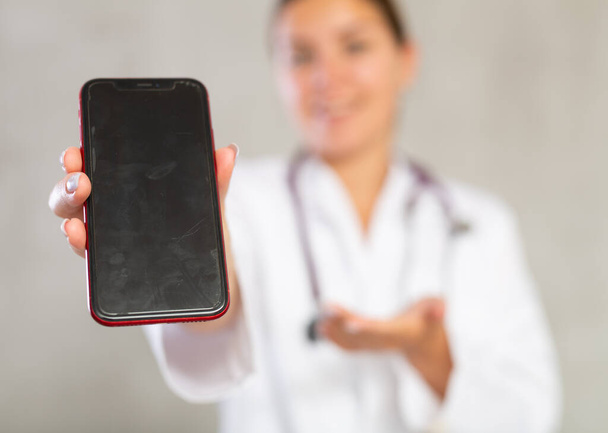 Female doctor in white coat extending hand with phone and showing screen, selective focus on gadget. Online consultation concept - Foto, Bild