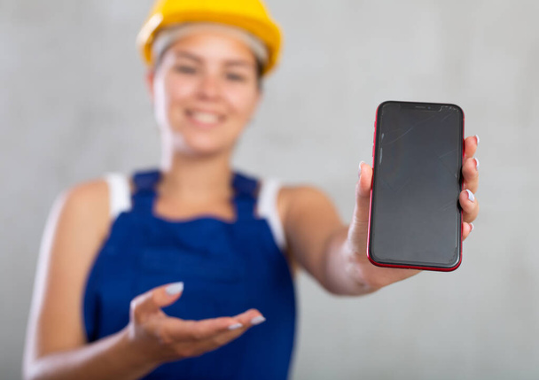 Closeup of professional smiling female engineer, construction manager in blue construction overalls and safety helmet, showing smartphone screen, grey background - 写真・画像