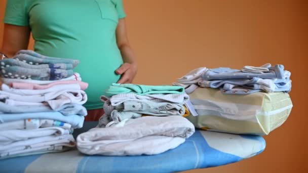 Middel van een zwangere vrouw, aanstaande moeder die haar buik strelt, aan de strijkplank staat met schone pasgeboren kleren, opvouwbare tas voor het zwangerschapsziekenhuis, zwanger wordt. Zwangerschap. Bevalling. - Video