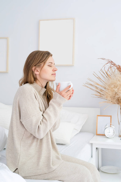caucasian woman waking up in her bed in the morning. young millennial woman sitting on the bed in the morning drinking coffee, light and airy room - Photo, Image