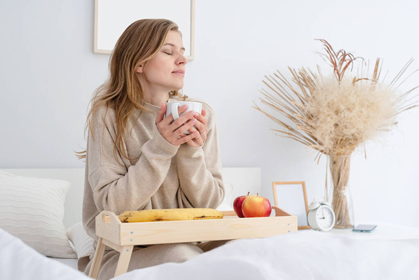 белая женщина просыпается в своей постели по утрам. молодая женщина тысячелетия сидит на кровати утром пить кофе, легкий и просторный номер - Фото, изображение