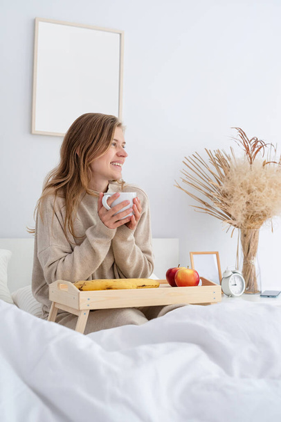 Biała kobieta budzi się rano w łóżku. młoda tysiącletnia kobieta siedzi rano na łóżku pijąc kawę, światło i przestronny pokój - Zdjęcie, obraz