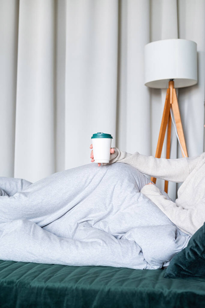 Kaukasische Frau wacht morgens in ihrem Bett auf. junge Millennial-Frau sitzt morgens auf dem Bett und trinkt Kaffee aus Pappbecher, Design-Attrappen - Foto, Bild