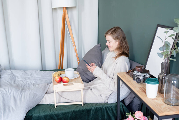 Kafkasyalı kadın sabahları yatağında uyanıyor. Genç milenyum kadını sabahları yatakta oturur kahve, ışık ve havadar oda içer. - Fotoğraf, Görsel