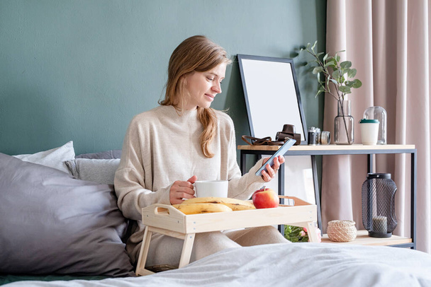 Biała kobieta budzi się rano w łóżku. młoda tysiącletnia kobieta siedzi rano na łóżku pijąc kawę, światło i przestronny pokój - Zdjęcie, obraz