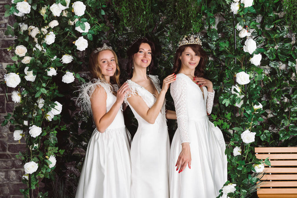 Trzy piękne panny młode w eleganckich białych sukienkach przed roślinami i łuk z kwiatami. Grupa narzeczonych w ogrodzie. - Zdjęcie, obraz