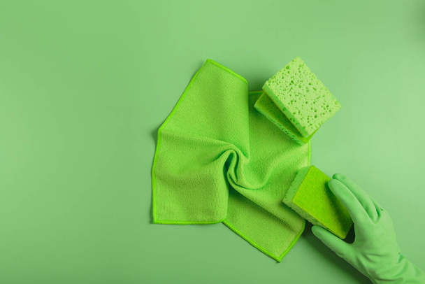 Cleaning set for various surfaces in kitchen, bathroom and other rooms on green background. Hand in rubber glove holding washing sponge. Cleaning concept, professional cleaning, household supplies - 写真・画像