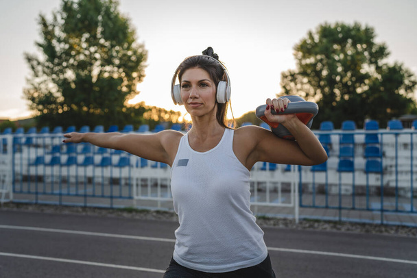 One woman adult caucasian female athlete training with kettlebell girya weight in stadium on track in summer evening or morning with headphones happy brunette health and fitness concept copy space - Fotó, kép