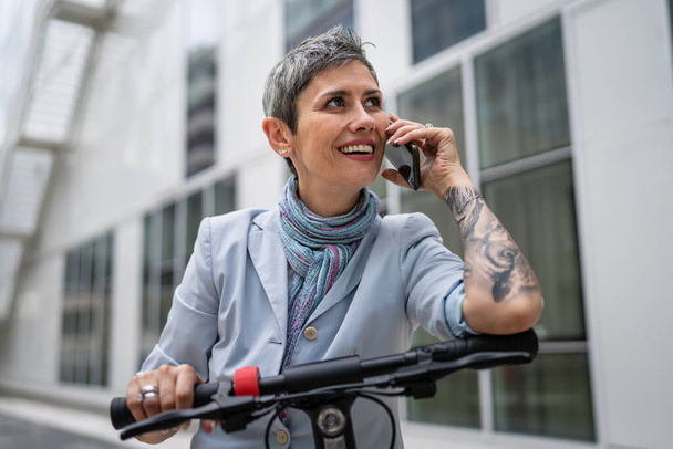 μία γυναίκα ώριμη ανώτερος καυκάσιος θηλυκό σταθεί υπαίθρια χρήση κινητό τηλέφωνο κάνοντας μια ομιλία κλήσης με ηλεκτρικό πατίνι την ημέρα αυτοπεποίθηση σύγχρονο εναλλακτικό τρόπο μεταφοράς και επικοινωνίας αντίγραφο χώρου - Φωτογραφία, εικόνα