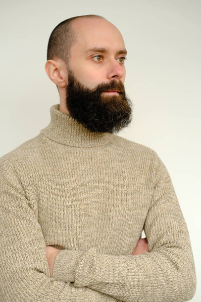 крупным планом мужское лицо, молодой задумчивый бородатый и усатый мужчина, довольный парень с 30 годами в бежевом шерстяном свитере серьезно отворачивается, концепция человеческого здоровья, размышления о смысле жизни - Фото, изображение
