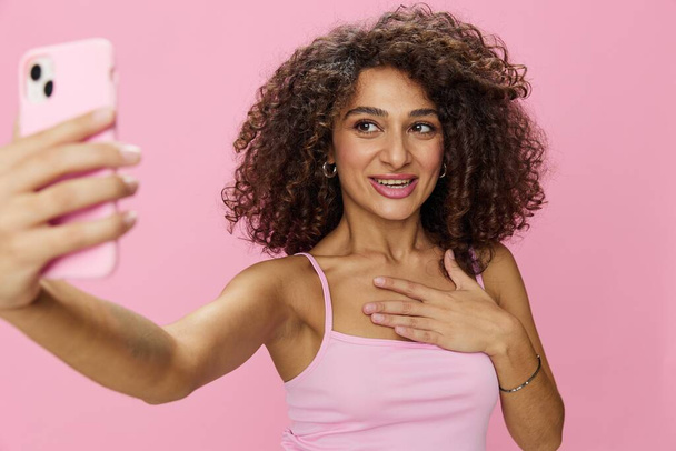 Bloggerin mit Telefonvideoanruf macht Selfies, mit lockigem Haar in rosa Lächeln T-Shirt und Jeans posiert auf rosa Hintergrund, Kopierraum, Technologie und soziale Medien, online. Hochwertiges Foto - Foto, Bild