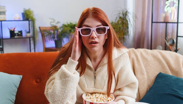 Mujer joven emocionada sentada en el sofá comiendo palomitas de maíz y viendo interesante serie de televisión, juego deportivo, película, contenido de películas en línea en redes sociales en casa. Chica en gafas 3D disfrutando del entretenimiento doméstico - Foto, imagen