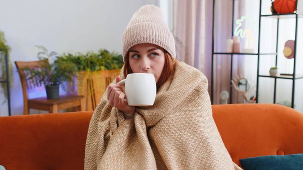 Άρρωστη κοκκινομάλλα φοράει καπέλο τυλιγμένο σε καρό κάθεται μόνη της τρέμοντας από το κρύο στον καναπέ πίνοντας ζεστό τσάι σε μη θερμαινόμενο διαμέρισμα χωρίς θέρμανση λόγω χρέους. Ανθυγιεινό κορίτσι αισθάνεται δυσφορία προσπαθούν να ζεσταθεί - Φωτογραφία, εικόνα