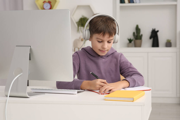 Boy writing in notepad while using computer and headphones at desk in room. Home workplace - Photo, Image