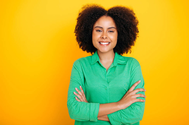 Portrait of a positive gorgeous hispanic or brazilian curly haired woman, wearing a green casual shirt, looking at the camera, smiling, standing with arms crossed on an isolated yellow background - Valokuva, kuva