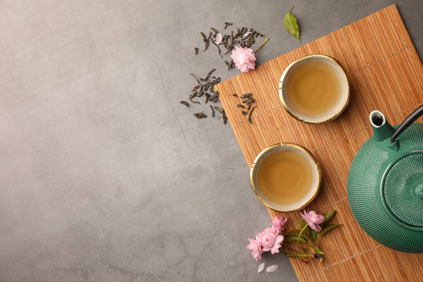 Traditional ceremony. Cups of brewed tea, teapot and sakura flowers on grey table, flat lay with space for text - Foto, Imagen