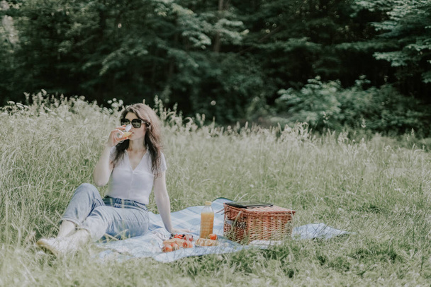 A beautiful young caucasian brunette girl in jeans, a t-shirt and sunglasses sits on a bedspread with a wicker basket, food and drinks orange juice from a glass in a public park, close-up side view. Outdoor picnic concept. - Photo, image